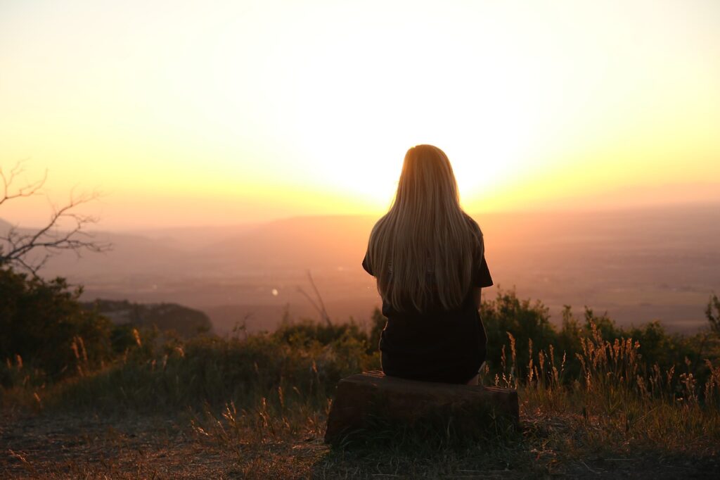 Mujer de espalda sentada en una piedra en el campo, en un lugar elevado, contemplando el amanecer o el ocaso con colores anaranjados